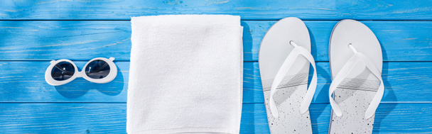 panoramic shot of white folded towel, retro sunglasses and flip flops on blue wooden background - Foto, imagen