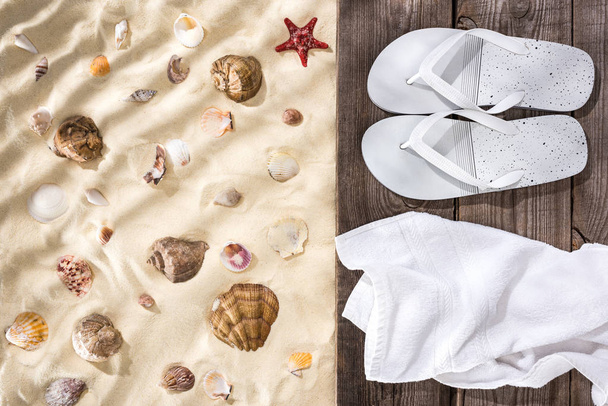 Draufsicht auf Muscheln und Seesterne auf Sand und Flip Flops und weißes Handtuch auf braunem Holzbrett - Foto, Bild