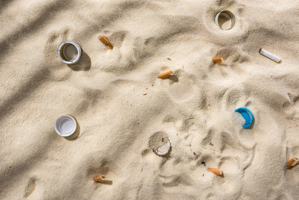 vista superior de conchas, tampas de garrafa, pontas de cigarro espalhadas, tampas de garrafa de plástico na areia
 - Foto, Imagem
