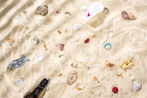 top view of seashells, scattered cigarette butts, apple core, plastic cups, glass bottle and candy wrapper on sand - Foto, Imagen