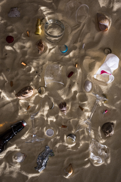 vista superior de conchas, garrafa de vidro, pontas de cigarro espalhadas, óculos quebrados, núcleo de maçã, copos de plástico e invólucro de doces na areia com sombras
 - Foto, Imagem