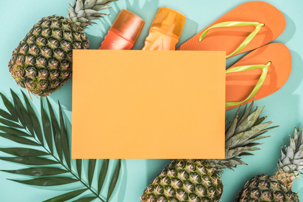 top view of pineapples, tropical leaf, sunscreens, orange flip flops and empty card on turquoise background - Photo, Image
