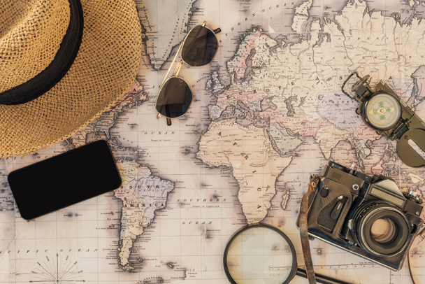 Pohled na slaměný klobouk, sluneční brýle, filmovou kameru, Lupa, kompas a smartphone s prázdnou obrazovkou na mapě světa - Fotografie, Obrázek