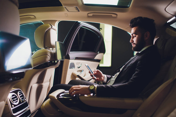 Día de trabajo ocupado. Hombre joven guapo en traje completo mirando su teléfono inteligente mientras está sentado en el coche
. - Foto, Imagen