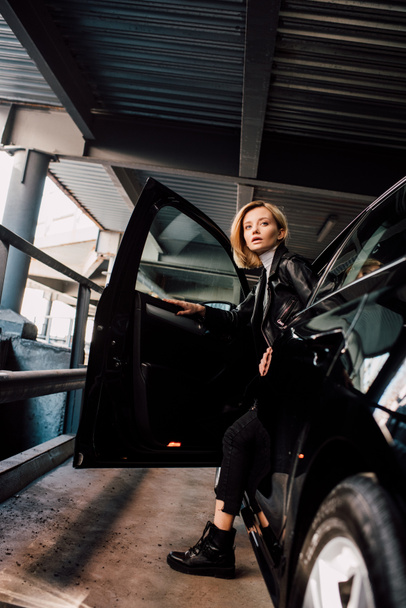 ελκυστική ξανθιά κορίτσι στέκεται κοντά στο μαύρο αυτοκίνητο με ανοιχτή πόρτα  - Φωτογραφία, εικόνα