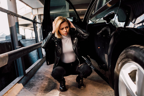 ελκυστική ξανθιά γυναίκα που έχει ημικρανία και κάθεται κοντά στο μαύρο αυτοκίνητο με ανοιχτή πόρτα  - Φωτογραφία, εικόνα