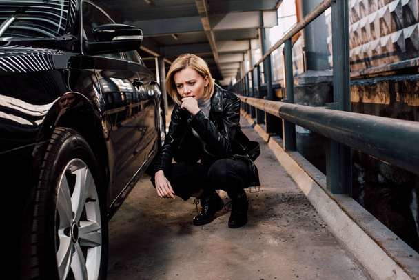 huolestunut blondi nainen istuu lähellä musta auto pysäköinti
  - Valokuva, kuva