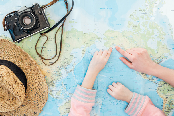 Vista recortada de niño y mujer con sombrero de paja y cámara de película apuntando con los dedos en el mapa del mundo
 - Foto, Imagen