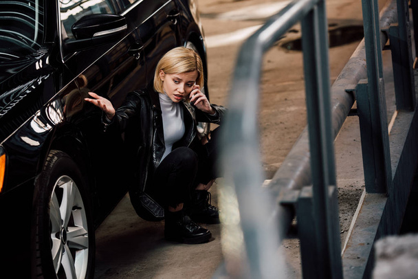 selective focus of sad blonde girl talking on smartphone while sitting near black car - Foto, Imagem