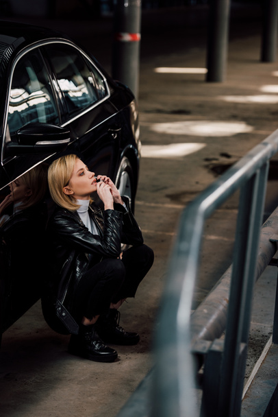 selective focus of worried blonde girl talking on smartphone while sitting near black car - Foto, Bild