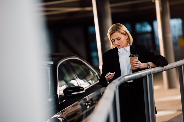selective focus of worried blonde girl holding smartphone and paper cup near black car - Photo, Image