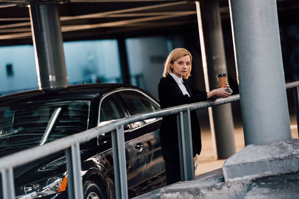 beautiful blonde girl holding smartphone and paper cup and standing near black car - Foto, Bild