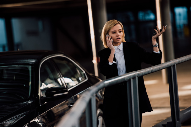 attractive blonde girl talking on smartphone and gesturing near black car - Fotografie, Obrázek