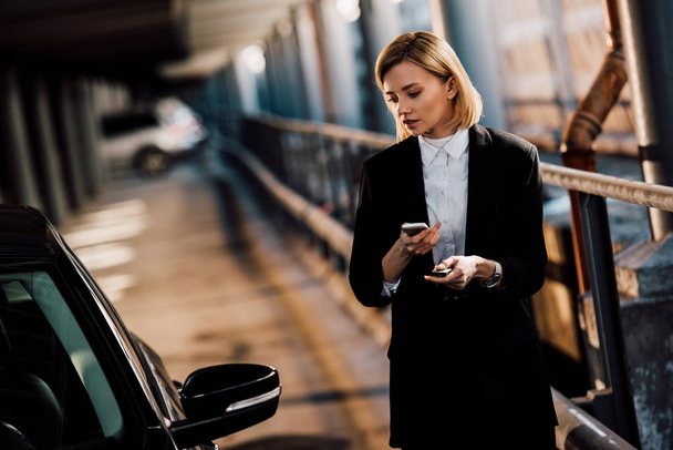 beautiful blonde girl holding smartphone and holding key near black car - Photo, Image