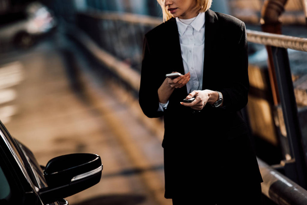 cropped view of blonde girl holding smartphone and key near black car - 写真・画像