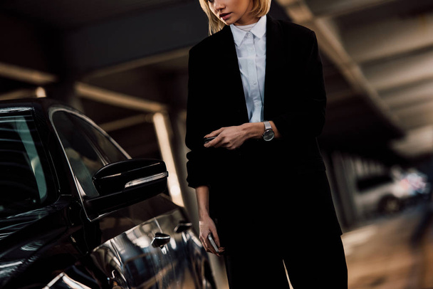 cropped view of blonde woman holding smartphone and key near black car - Фото, изображение