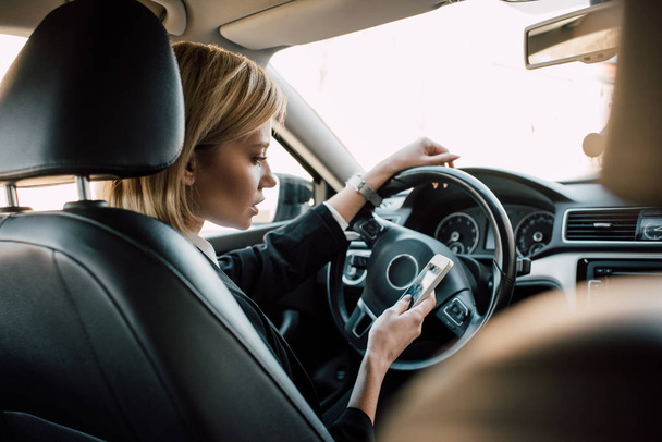 attractive blonde woman sitting in car and using smartphone  - Foto, afbeelding