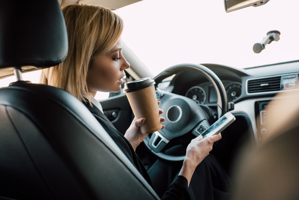 blonde young woman holding paper cup while looking at smartphone in car  - Valokuva, kuva