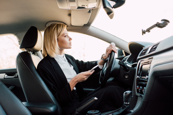 attraente bionda giovane conducente in possesso di smartphone durante la guida auto
  - Foto, immagini