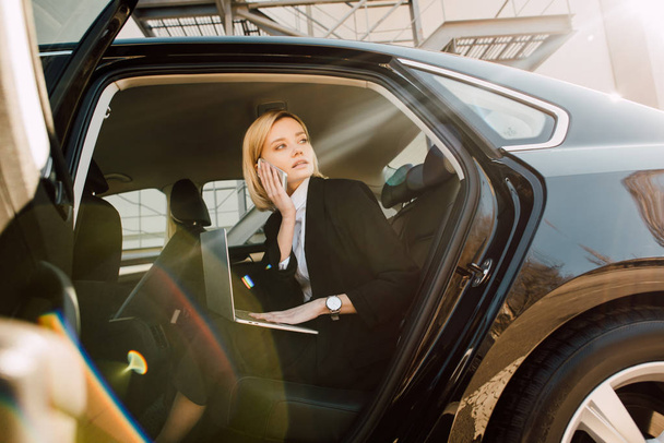 low angle view of blonde woman talking on smartphone while sitting with laptop in car  - Foto, immagini