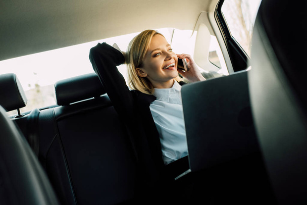 happy blonde woman talking on smartphone and smiling near laptop in car  - Fotoğraf, Görsel