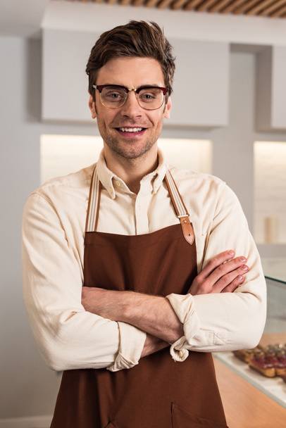 Smiling barista in glasses and brown apron looking at camera with crossed arms - Fotografie, Obrázek
