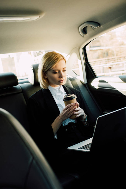 attractive blonde girl holding paper cup and looking at laptop in car  - Фото, изображение