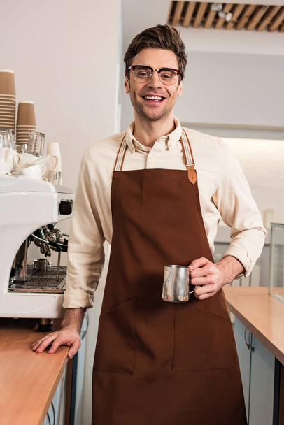 Kafede süt sürahi tutan gözlük ve kahverengi önlük gülümseyen barista - Fotoğraf, Görsel