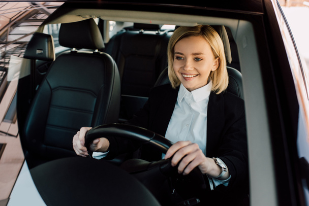 cheerful blonde driver holding steering wheel while driving car  - Fotó, kép