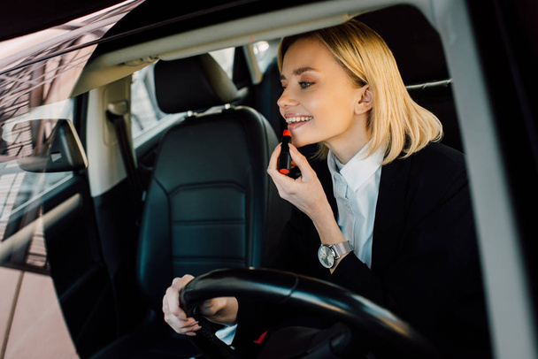 cheerful blonde young woman applying lipstick in car  - Фото, зображення