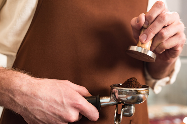 Cropped view of barista in brown apron holding portafilter with ground coffee and tamper - Fotó, kép