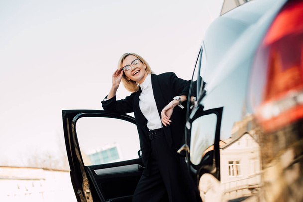 low angle view of happy blonde young woman in glasses standing near black automobile and touching glasses  - Photo, image