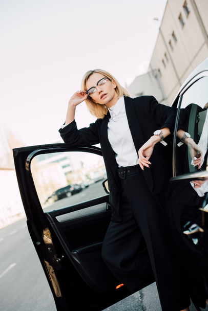 serious blonde young woman in glasses standing near black automobile and touching glasses   - Φωτογραφία, εικόνα