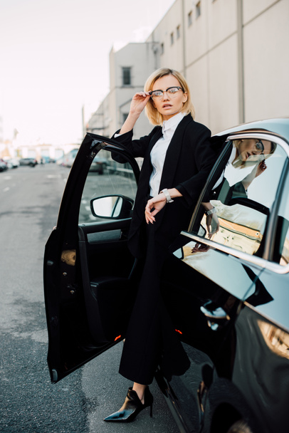 blonde young woman in glasses standing near black automobile and touching glasses   - Zdjęcie, obraz