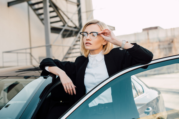 confident blonde young woman in glasses standing near black automobile and touching glasses   - Photo, image