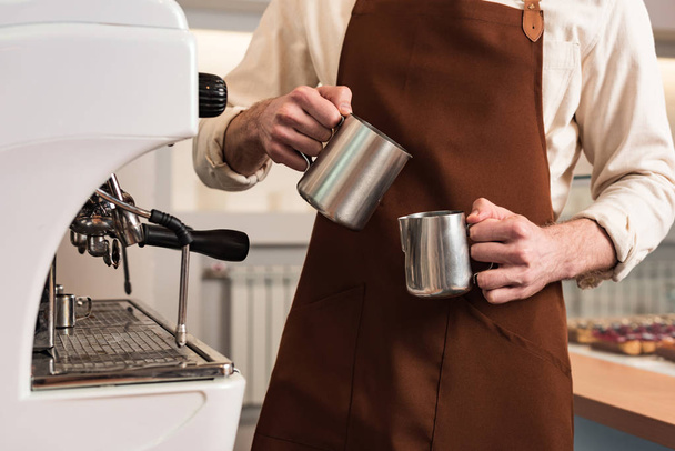 Cropped view of barista in brown apron holding steel jugs - Photo, Image
