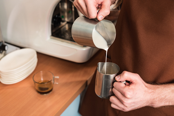 Cropped view of barista in brown apron pouring milk in steel jug - Fotó, kép