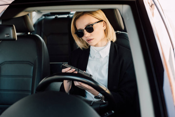 attractive young blonde woman in sunglasses holding gun in car  - Fotografie, Obrázek