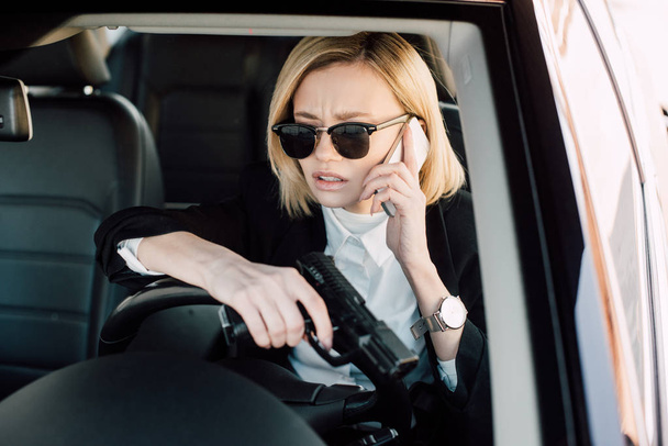 blond kobieta w okulary rozmawiać na smartphone i trzymając pistolet w samochodzie  - Zdjęcie, obraz