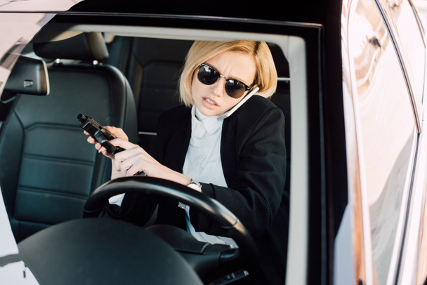 confident young blonde woman in sunglasses talking on smartphone and holding gun in car  - Φωτογραφία, εικόνα