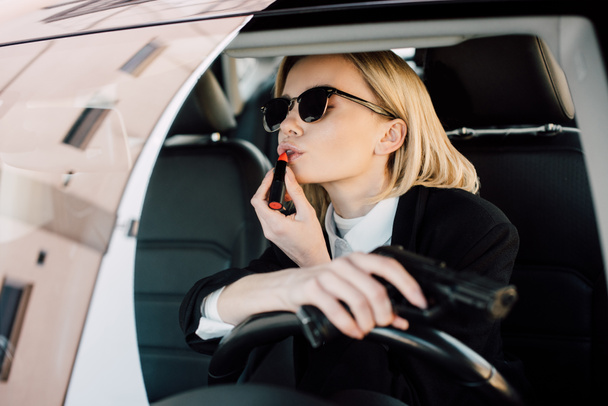 attractive blonde woman applying lipstick while holding gun in car  - Фото, зображення
