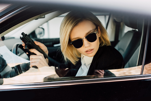 serious young blonde woman in sunglasses holding gun in car  - Fotografie, Obrázek