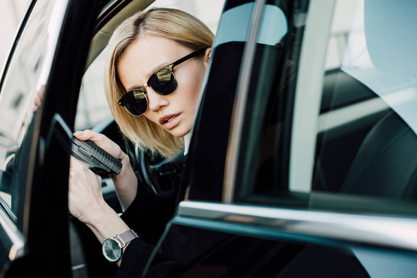 serious blonde woman in sunglasses holding gun in car  - Фото, изображение
