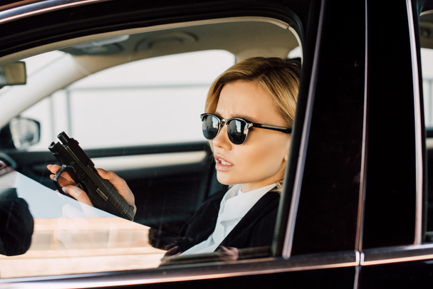 confident young blonde woman in sunglasses holding gun in car  - 写真・画像