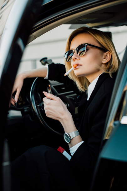 blonde woman in sunglasses holding lighter while smoking cigarette in car  - Фото, изображение