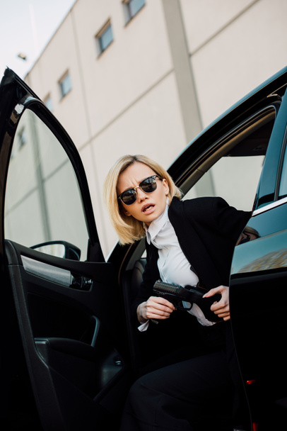 emotional blonde woman in sunglasses holding gun near black car  - Фото, изображение