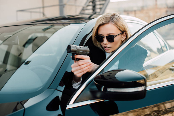 confident blonde woman in sunglasses holding gun near black car  - Foto, Imagem