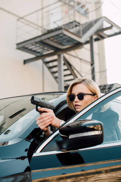 serious blonde woman in sunglasses holding gun near black car  - Fotografie, Obrázek
