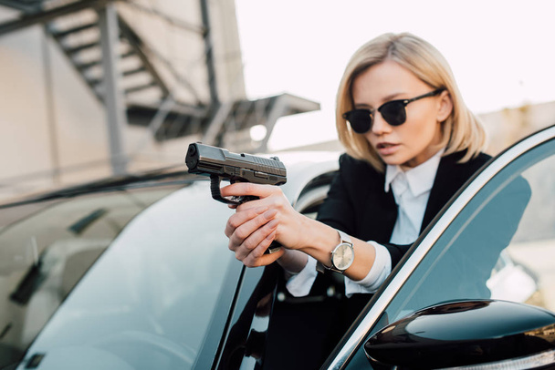 selective focus of serious blonde woman in sunglasses holding gun near black car  - Φωτογραφία, εικόνα