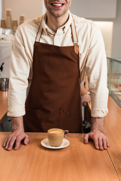 Обрізаний вид сміється бариста в коричневому фартуху з чашкою кави
 - Фото, зображення
